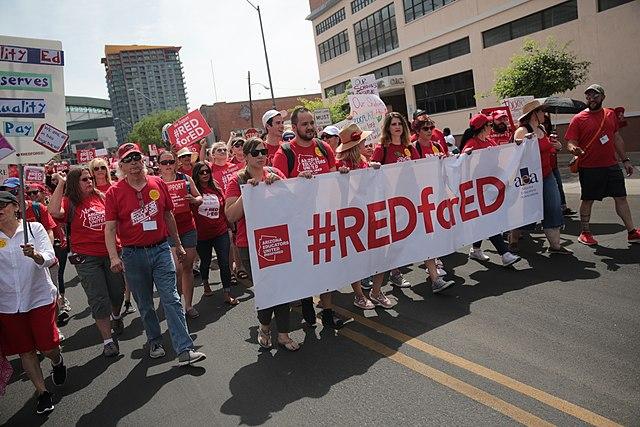 A large crowd of teachers, all wearing red, march behind a banner that reads &quot;Red for Ed.&quot;