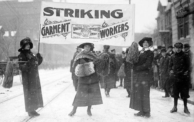 A black and white image of three women holding a banner which reads &quot;striking garment workers&quot; in front of a crowd of striking workers on a snowy day.